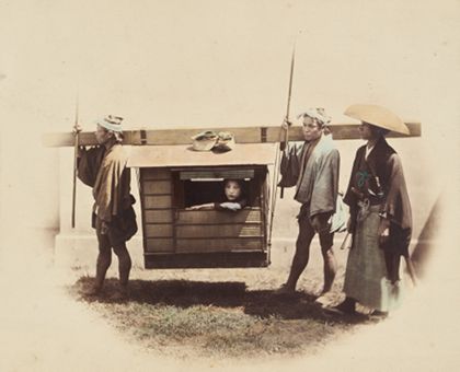 Fotografie, die eine Frau in einer Sänfte zeigt. Die Sänfte wird von zwei Männern getragen und von einem Mann mit zwei Schwertern, der als Samurai posiert, bewacht.