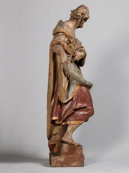 Stehende Holzskulptur des Apostels Simon Zelotes in theatralisch verdrehter Körperhaltung und ebenso expressiver Drapierung des stoffreichen Gewands, Seitenansicht von links.