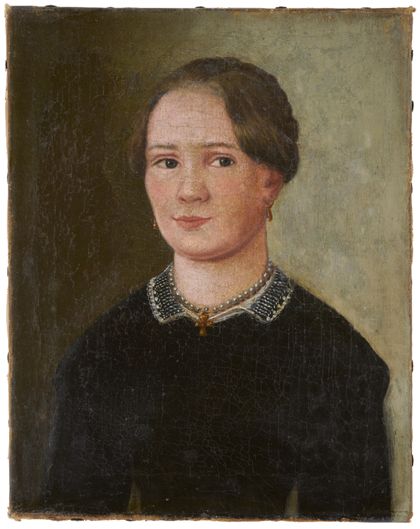 Ausgerahmtes Porträt einer jungen Frau, schwarz gekleidet, Perlenkette mit goldenem Kreuzanhänger und Ohrringen. Ihre Augen blicken zur Seite.