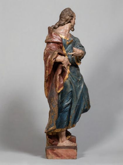 Stehende Holzskulptur des Apostels Judas Thaddäus in theatralisch verdrehter Körperhaltung und ebenso expressiver Drapierung des stoffreichen Gewands, Seitenansicht von links.
