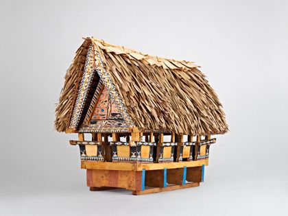 Hölzernes Modell eines Versammlungshauses. Das Haus steht auf einem Sockel aus vier großen Balken. Gebälk und Giebel-Verschalung sind beschnitzt und mehrfarbig mit menschlichen und tierischen Figuren bemalt. Das Dach ist mit Blattwerk gedeckt.